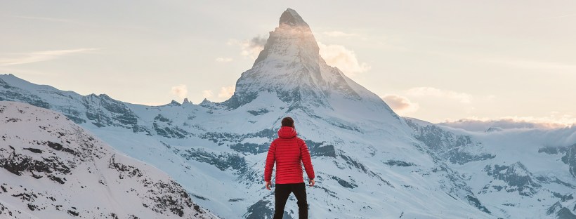 A man in a red parka stands on a snow covered hill looking at jagged mountain peak ahead of him